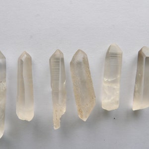 May include: Six clear quartz crystal points, also known as wands, are arranged in a row on a white surface. The points are all different sizes and shapes, but they are all clear and have a natural, unpolished finish.