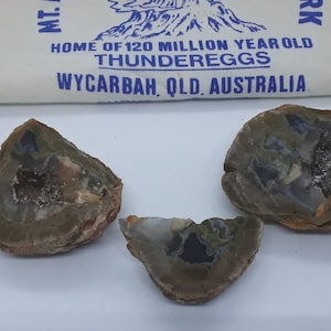 May include: Three polished thunder eggs in shades of brown, grey, and blue. The cut stones are displayed with a white cloth bag that has text that reads "HOME OF 120 MILLION YEAR OLD THUNDEREGGS WYCARBAH, QLD, AUSTRALIA."