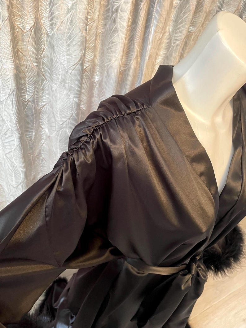 May include: A black satin robe with long sleeves and a wrap-around design. The sleeves are gathered at the shoulders, and the robe is trimmed with black feathers. A matching belt is tied at the waist.