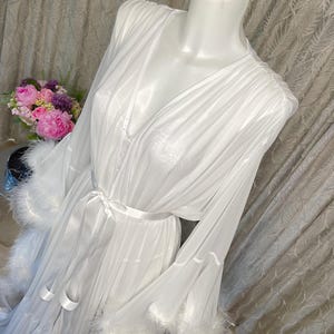Puede incluir: Una bata blanca y transparente con mangas largas y cintura anudada. Las mangas y el dobladillo est&aacute;n adornados con plumas blancas y esponjosas. La bata se exhibe en un maniqu&iacute;, con un ramo de flores rosas y moradas en el fondo.