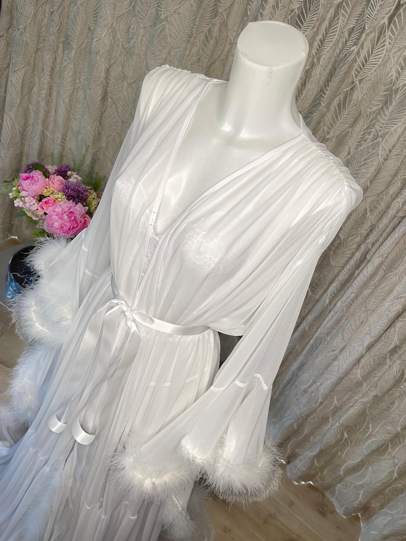 Puede incluir: Una bata blanca y transparente con mangas largas y cintura anudada. Las mangas y el dobladillo est&aacute;n adornados con plumas blancas y esponjosas. La bata se exhibe en un maniqu&iacute;, con un ramo de flores rosas y moradas en el fondo.