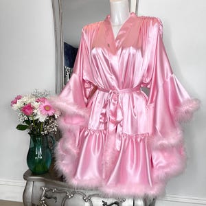 May include: A pink satin robe with a ruffled hem and feather trim. The robe has long sleeves with a tie at the waist.