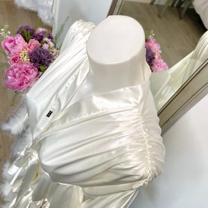 May include: Ivory satin robe with feather trim, displayed on a mannequin. The robe features a draped design with gathered fabric on the sleeves. A small label is visible on the robe. A bouquet of pink and purple flowers is in the background.