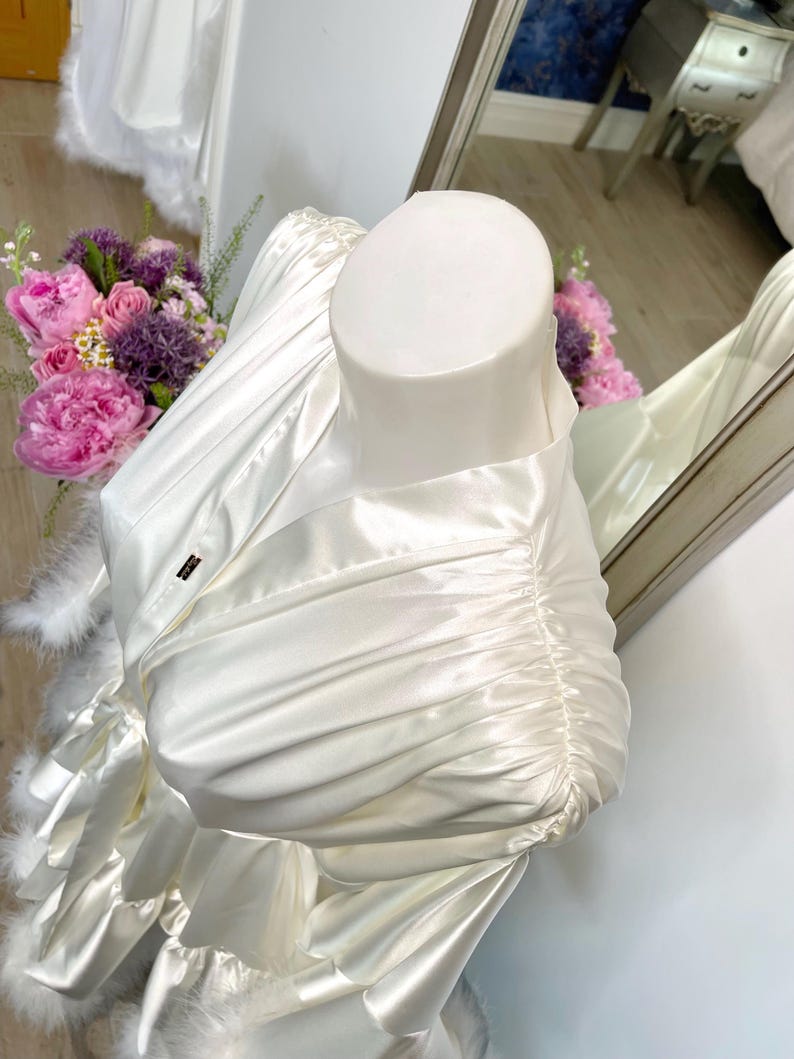 May include: Ivory satin robe with feather trim, displayed on a mannequin. The robe features a draped design with gathered fabric on the sleeves. A small label is visible on the robe. A bouquet of pink and purple flowers is in the background.