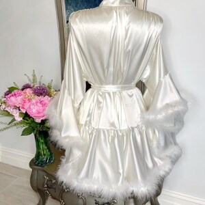 May include: A white satin robe with a matching belt and feather trim on the sleeves and hem. The robe is displayed on a mannequin, with a vase of pink flowers and a mirror in the background.