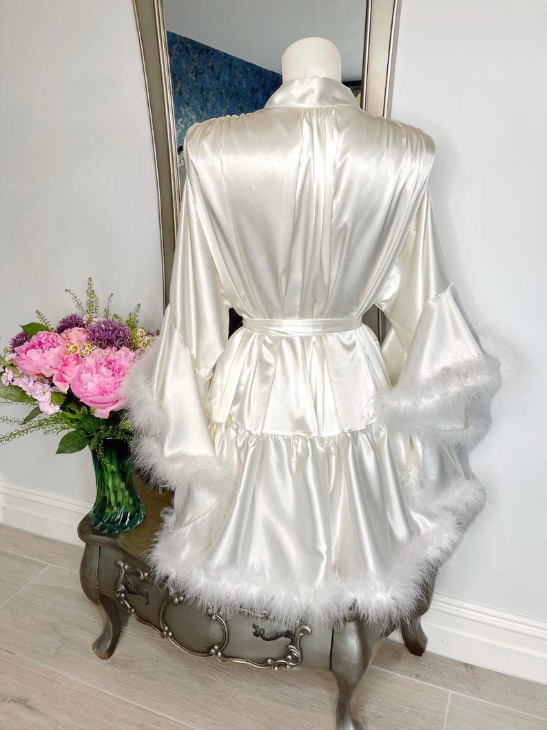 May include: A white satin robe with a matching belt and feather trim on the sleeves and hem. The robe is displayed on a mannequin, with a vase of pink flowers and a mirror in the background.