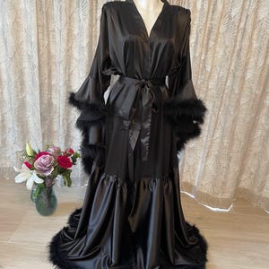 Może przedstawiać: Czarny satynowy szlafrok z pasującym paskiem i obszyciem z piór. Szlafrok ma długie rękawy z mankietami z piór i falbanką u dołu. Szlafrok jest prezentowany na manekinie.