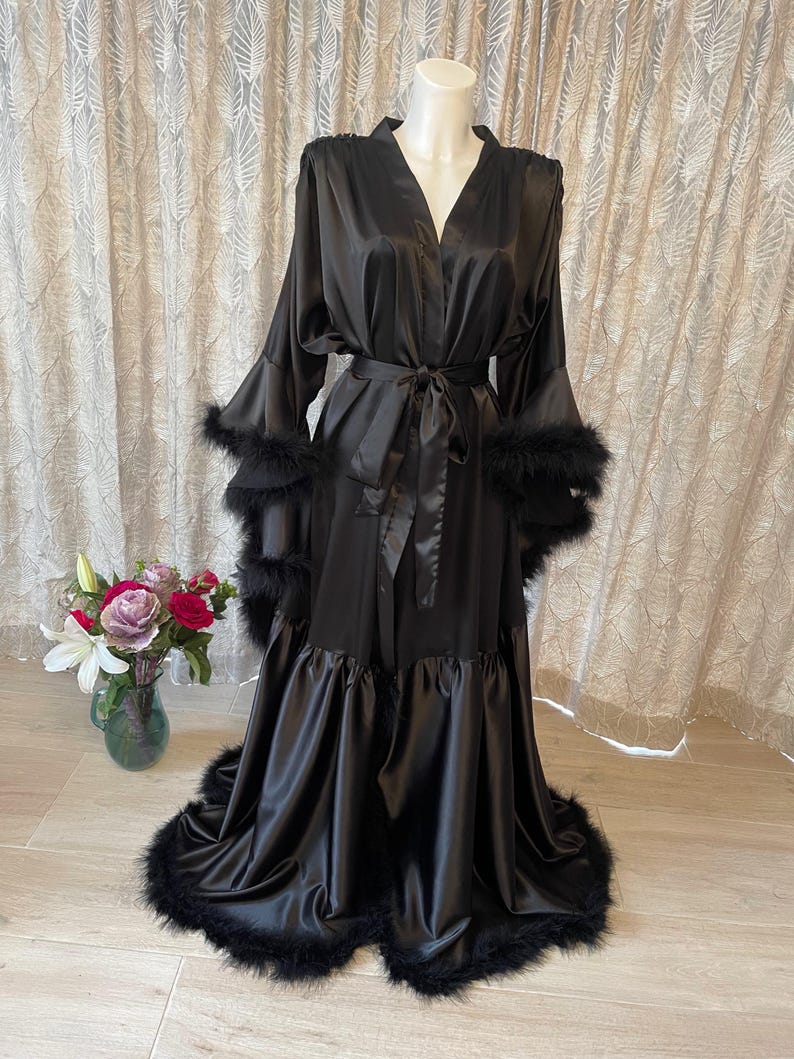 May include: A black satin robe with a matching belt and feather trim. The robe has long sleeves with feather cuffs and a ruffled hemline. The robe is displayed on a mannequin and is set against a neutral background.