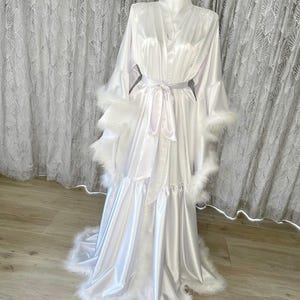 May include: A white satin robe with a ruffled hem and feather trim. The robe has a sash tie at the waist and long sleeves.