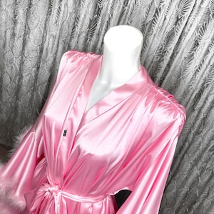May include: A pink satin robe with long sleeves and a tie belt. The sleeves have white feather trim.