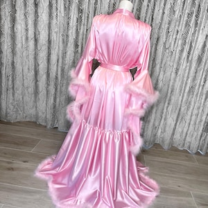 May include: A pink satin robe with a ruffled hem and feather trim. The robe has long sleeves with bell cuffs and a sash tie at the waist.