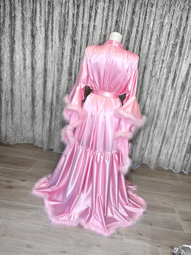 May include: A pink satin robe with a ruffled hem and feather trim. The robe has long sleeves with bell cuffs and a sash tie at the waist.