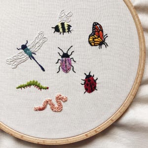 May include: Embroidered hoop art featuring a variety of insects, including a dragonfly, a bee, a butterfly, a ladybird, and a worm. The insects are embroidered in various colours on a white fabric background.