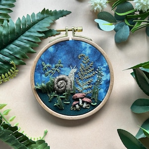 May include: An embroidered artwork in a wooden hoop features a snail, mushrooms, and foliage on a blue and green background. The embroidery is detailed with various textures and colours, creating a miniature woodland scene. The hoop is approximately 15 cm in diameter.
