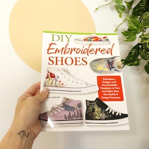 May include: A book titled "DIY Embroidered Shoes" with a green background and a white circle. The book cover features several pairs of shoes with different embroidery designs, including a pair of white shoes with red birds and flowers, a pair of purple shoes with a forest scene, and a pair of black shoes with a floral design. The book cover also includes the text "Techniques, Designs, and Downloadable Templates to Turn Any Fabric Shoe into Stylish & Unique Footwear."