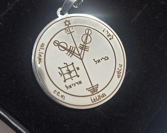 May include: A silver-colored pendant necklace with a circular charm featuring engraved symbols and text. The pendant hangs on a matching chain, presented in a black box. The design includes a six-pointed star and other intricate patterns.
