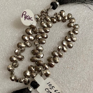 May include: Strands of faceted pyrite beads with a metallic silver color. The beads are teardrop-shaped and strung together. Black tassels are attached at both ends. The image includes a tag that reads "Pyrite".