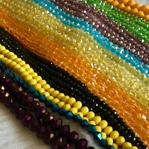 May include: A close-up of multiple strands of colorful beads. The beads are arranged in parallel rows, showcasing a variety of colors including yellow, orange, green, blue, purple, and black. The beads have a faceted, crystal-like appearance.