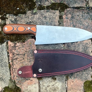 May include: A fixed-blade knife with a silver-colored metal blade and a handle featuring alternating orange and black layered patterns. The knife is accompanied by a dark brown leather sheath with red stitching and a silver metal snap closure.