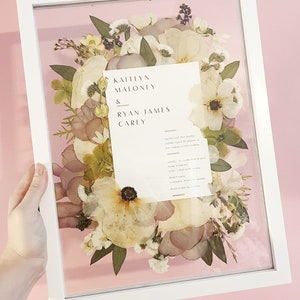 May include: A framed floral arrangement featuring dried flowers in shades of white, cream, and pale pink. A white rectangular frame surrounds the flowers and a card with the names "Kaitlyn Maloney & Ryan James Carey" is visible. The background is a soft pink.