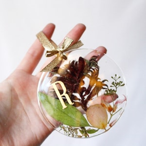 May include: A clear glass ornament with a gold ribbon and a gold letter "R" inside. The ornament is filled with dried flowers and leaves in shades of brown, green, and pink.