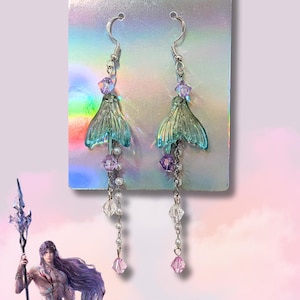 May include: A pair of silver-toned mermaid tail earrings. The earrings feature a teal-colored tail design, accented with purple and clear crystal beads, and small white pearls. The earrings hang from a silver hook.
