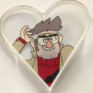 Op de afbeelding: Een hartvormige acrylpin met een cartoonfiguur met bruin haar, een rode trui en een bruine jas. Het personage glimlacht en heeft een ondeugende blik in zijn ogen.