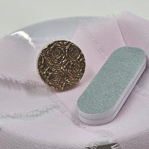May include: A round, gold-colored stamp with a rose pattern next to a gray nail file. The items rest on a light pink fabric. The stamp has a detailed floral design, and the nail file has a rectangular shape with white edges.