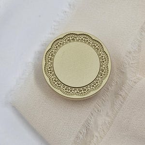May include: A round, gold-colored stamp with a detailed lace-like border. The stamp has a blank center and is resting on a piece of light beige fabric with a frayed edge. The background is a soft, neutral color.