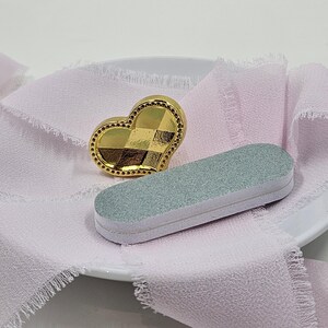 May include: A gold heart-shaped object with a checkered design and a small, light green nail file on a white plate with pink fabric. The heart has a beaded border.