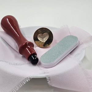 May include: A burgundy wax seal stamp with a black tip, a gold heart-shaped seal, and a gray nail file rest on a pink fabric. The items are arranged on a white plate, suggesting a crafting or stationery theme.