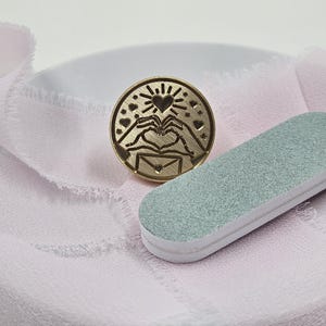 May include: A round, gold-colored wax seal stamp with a heart and hands design, a sunburst, and small hearts. A light green and white nail file rests beside the stamp on a pink fabric.
