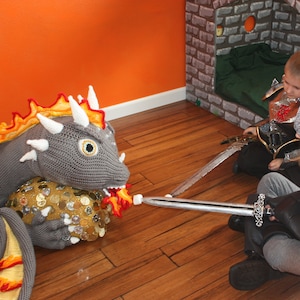 Può includere: Un drago grigio all'uncinetto con fiamme arancioni e gialle sulla schiena è rivolto a un bambino vestito da cavaliere. Il cavaliere tiene una spada d'argento ed è seduto su un pavimento di legno.