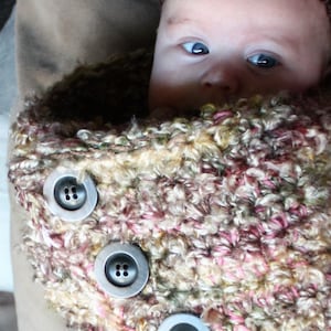 May include: A baby wearing a crocheted, multi-colored,  knitted baby wrap with three buttons. The wrap is a mix of pink, white, and brown yarn.