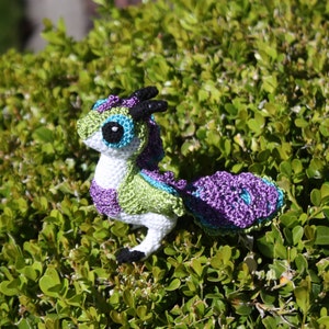 Puede incluir: Un juguete de dragón de crochet hecho con hilo verde, morado y blanco. El dragón tiene una pequeña cara blanca con ojos negros y una cola morada.