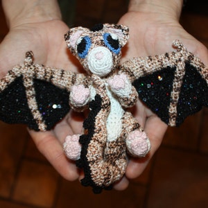 Puede incluir: Un dragón de peluche tejido a crochet con pelaje marrón y blanco, alas negras con purpurina y ojos azules.