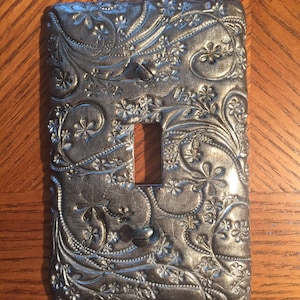 Denna eleganta och vackra strömbrytarplatta har ett blommigt vinmönster i en skimrande pistolmetallfärg. Enkla vippbara ljusbrytarplattor. Stora beställningar.