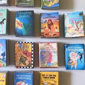 May include: A collection of miniature children's books featuring classic stories like 'The Lion King', 'Sleeping Beauty', 'Little Red Riding Hood', and 'The Real Mother Goose'. The books are arranged in a grid pattern on a white surface.