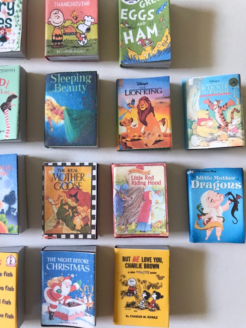 May include: A collection of miniature children's books featuring classic stories like 'The Lion King', 'Sleeping Beauty', 'Little Red Riding Hood', and 'The Real Mother Goose'. The books are arranged in a grid pattern on a white surface.