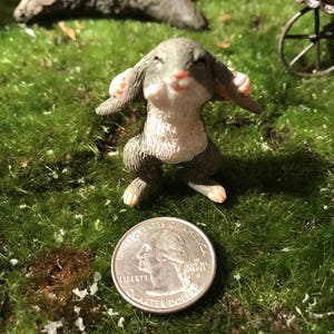 May include: A small gray and white rabbit figurine with its ears covering its eyes. The figurine is standing on a bed of green moss.