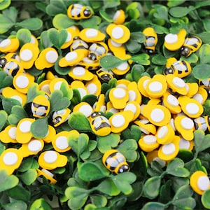 May include: A large quantity of yellow and black wooden bee-shaped embellishments with white adhesive backing. The bees are scattered on a bed of green artificial grass.