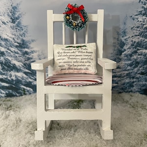 Op de afbeelding: Een witte schommelstoel met een rood-wit gestreept kussen. Het kussen heeft een wit kussen met de tekst "Navidad en el Cielo, Que hacen ahi? Ellos bajan del cielo para pasar tiempo contigo. Entonces, guárdeles un asiento, solo una silla vacía. No los podrás ver, pero ellos estarán alli." Een groene en zilveren kerstkrans met een rode strik hangt aan de rugleuning van de stoel.