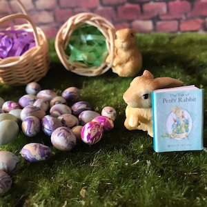 May include: A miniature scene with a wicker basket filled with purple ribbon, a bunny figurine reading a book titled "The Tale of Peter Rabbit", and a collection of colorful Easter eggs.