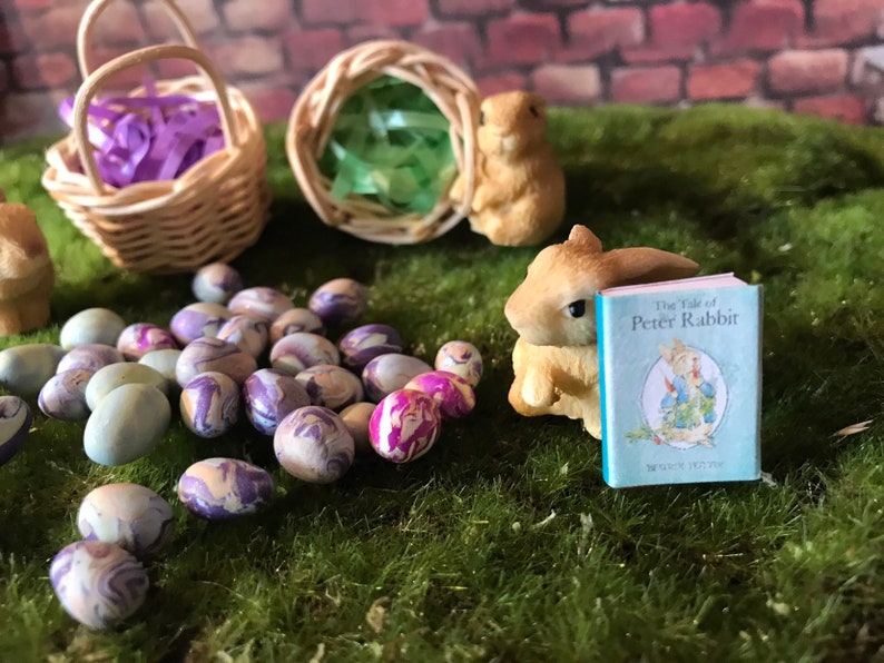May include: A miniature scene with a wicker basket filled with purple ribbon, a bunny figurine reading a book titled "The Tale of Peter Rabbit", and a collection of colorful Easter eggs.