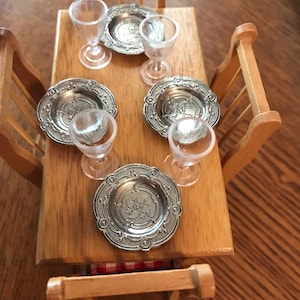 May include: A miniature wooden table set with four chairs, four silver-colored plates, and four clear glass wine glasses.