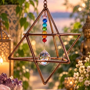 May include: A copper-colored geometric star-shaped frame hanging from a chain. Inside the frame, a vertical strand of seven small, multi-colored beads in rainbow order hangs above a single, clear, faceted crystal sphere that reflects light.
