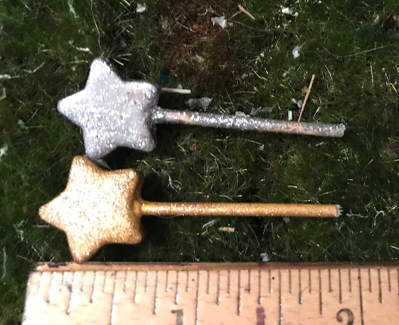 May include: Two miniature star-shaped wands, one silver and one gold, with glitter accents. The wands are approximately 2 inches long and are shown next to a ruler.