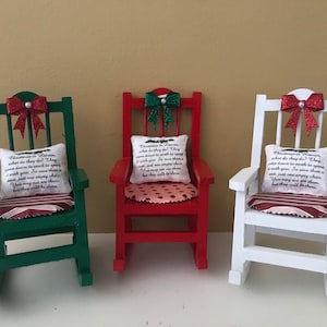 May include: Three miniature rocking chairs decorated for Christmas. The chairs are painted green, red, and white. Each chair has a red bow with glitter, a cushion with a Christmas-themed message, and a striped seat cushion.