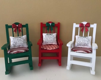 May include: Three miniature rocking chairs decorated for Christmas. The chairs are painted green, red, and white. Each chair has a red bow with glitter, a pillow with a Christmas-themed message, and a striped seat cushion.