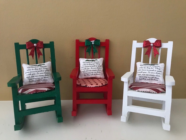 May include: Three miniature rocking chairs decorated for Christmas. The chairs are painted green, red, and white. Each chair has a red bow with glitter, a cushion with a Christmas-themed message, and a striped seat cushion.